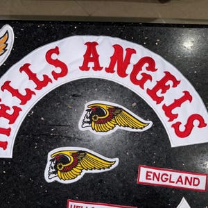 Puede incluir: Parches bordados con las palabras "HELLS ANGELS" en rojo, arqueadas sobre un fondo blanco. Los parches incluyen una calavera con alas y un parche rectangular que dice "ENGLAND".