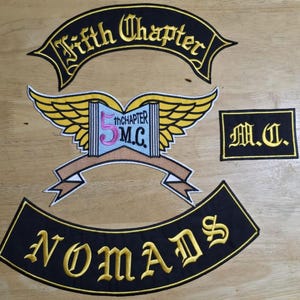 Op de afbeelding: Geborduurde patches met een motorclubthema. De patches bevatten de woorden "Fifth Chapter", "Nomads" en "M.C.". De centrale patch toont een boek met vleugels en het nummer "5". De patches zijn zwart met gouden en witte accenten.