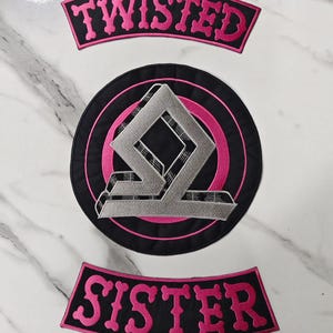 May include: Embroidered patches with the words "TWISTED" and "SISTER" in pink lettering on black fabric. A circular patch in the center features a geometric design in gray and black, outlined in pink.