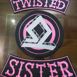 Puede incluir: Conjunto de tres parches bordados. El parche superior dice "TWISTED" en rosa sobre fondo negro. El parche central es un círculo negro con un anillo rosa y un diseño geométrico plateado y blanco. El parche inferior dice "SISTER" en rosa.