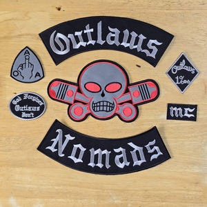 May include: A collection of black and gray embroidered patches with white lettering. The patches feature skull and piston designs, along with the words "Outlaws," "Nomads," and "God Forgives Outlaws Don't." A patch displays a hand gesture.