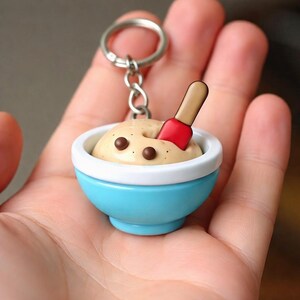 Op de afbeelding: Een miniatuur sleutelhanger met een lichtblauwe kom met een witte rand. Binnenin zit een bruinachtige substantie met twee bruine chocoladechip-achtige details en een rode spatel. Een zilveren sleutelring is bevestigd.