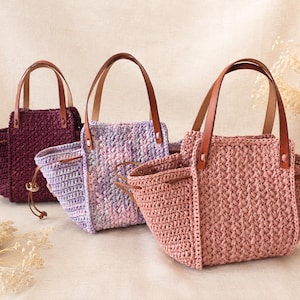 May include: Three crochet handbags with brown leather handles and drawstring closures. The bags are in deep purple, variegated lavender and white, and dusty rose colors. Each bag features a textured stitch pattern and a wide, open-top design.