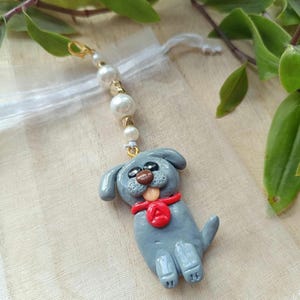 Könnte beinhalten: Ein handgefertigter grauer Hundeanhänger mit rotem Schal und goldenem Verschluss. Der Anhänger ist an einer Kette aus Kunstperlen und goldenen Perlen befestigt. Der Hund hat eine braune Nase und eine rote Zunge. Der Anhänger ist ca. 5 cm lang.