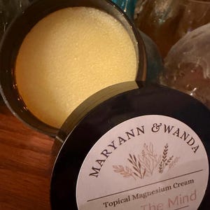 May include: A black jar of topical magnesium cream, with the lid partially open. The cream inside is a pale yellow. The lid is labelled with the words "Maryann & Wanda" and "Topical Magnesium Cream".