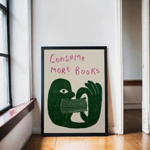 Könnte beinhalten: Ein gerahmter Kunstdruck mit den Worten "Consume More Books" in Rosa. Das Kunstwerk zeigt eine grüne Figur, die einen Stapel Bücher konsumiert. Der Druck wird vor einem neutralen Hintergrund an einer Wand gezeigt.
