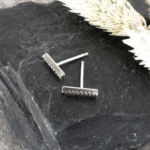 May include: Two silver stud earrings with a textured bar design. The earrings are on a dark gray stone surface.