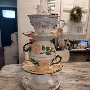 May include: A stacked teacup sculpture featuring a variety of vintage teacups and saucers. The teacups are white with floral designs in pink, yellow, and blue. The sculpture is topped with a small teacup and a white candle.