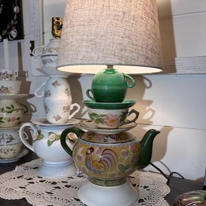 May include: A unique lamp crafted from stacked teacups, saucers, and a teapot, topped with a gray fabric lampshade. The teapot base features a rooster design. The lamp sits on a white lace doily.