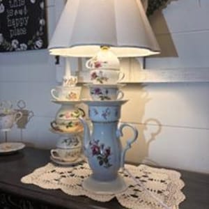 May include: A unique lamp made from stacked teacups and a teapot, topped with a white lampshade. The lamp features floral designs and sits on a decorative doily. The text on the sign in the background reads "this is our happy place."