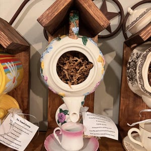 May include: A whimsical teapot birdhouse with a floral design, perched on a wooden stand. A small pink teacup and saucer sit below the spout. The birdhouse is filled with brown packing material. Other decorative teacups are visible.