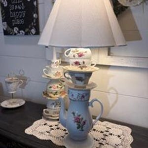May include: A unique lamp featuring a white lampshade atop a stack of vintage teacups, saucers, and a teapot. The teapot and teacups are adorned with floral patterns. The lamp sits on a dark table with a lace doily, with a sign that reads "this is our happy place".