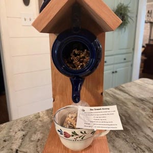 May include: A wooden bird feeder with a blue teapot-shaped container for birdseed. A white teacup with floral designs holds a birdseed treat. The feeder has a small roof and is mounted on a wooden base.