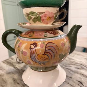 May include: A stacked ceramic teapot with a rooster design, topped with a teacup, saucer, and another teacup and saucer. The teapot is yellow with a rooster illustration and pink floral accents. The teacups and saucers are green and cream-colored.