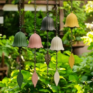 May include: Six bell-shaped wind chimes in shades of green, pink, black, white, and yellow. Each chime hangs from a chain and has a leaf-shaped accent. The chimes are suspended outdoors against a backdrop of green foliage, creating a tranquil garden scene.
