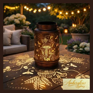 May include: A bronze-colored metal lantern with a handle, featuring cut-out mushroom and floral designs. The lantern casts a warm, patterned glow onto the wooden table. The image includes the text "Amberglow Lighting".