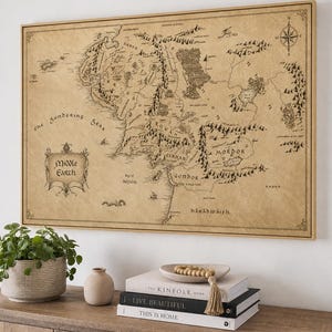 Può includere: Una mappa da parete rettangolare incorniciata della Terra di Mezzo stampata su carta color marrone chiaro in stile pergamena. La mappa presenta illustrazioni a inchiostro nero di montagne, foreste e coste, con il testo Middle Earth in una cornice.