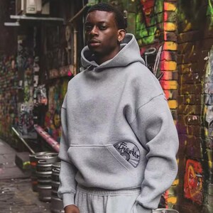 Puede incluir: Una persona con una sudadera con capucha gris y un pantal&oacute;n de ch&aacute;ndal a juego. La sudadera tiene un bolsillo delantero con un parche circular. El fondo presenta una pared cubierta de grafitis y barriles de metal.