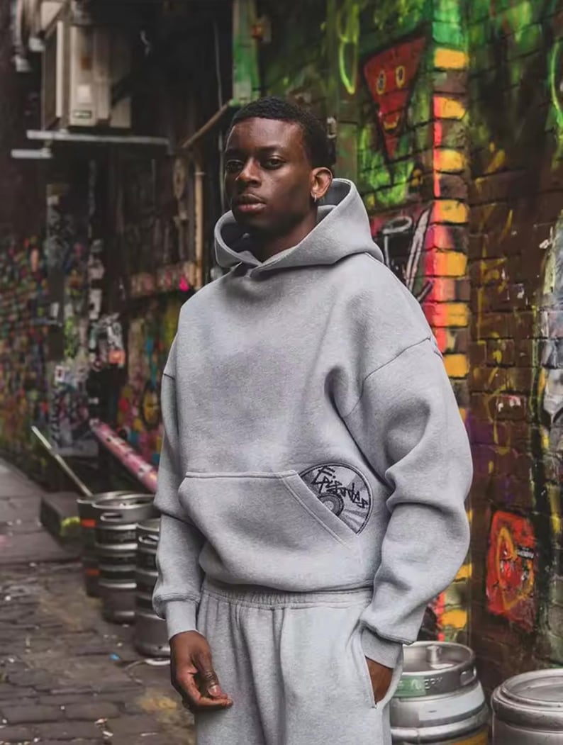 Puede incluir: Una persona con una sudadera con capucha gris y un pantal&oacute;n de ch&aacute;ndal a juego. La sudadera tiene un bolsillo delantero con un parche circular. El fondo presenta una pared cubierta de grafitis y barriles de metal.