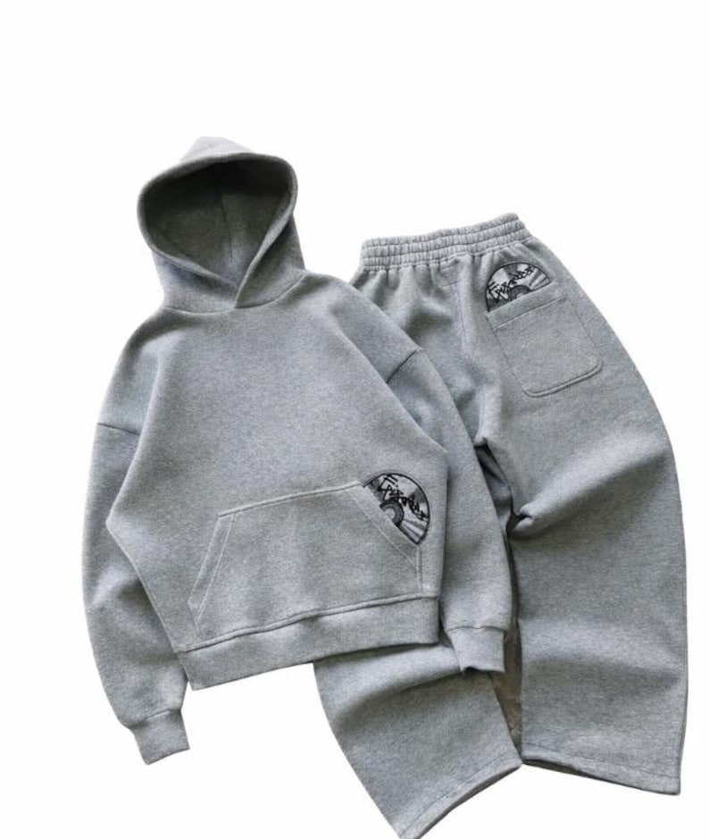 Puede incluir: Un conjunto de sudadera con capucha y pantal&oacute;n de ch&aacute;ndal grises. La sudadera tiene un bolsillo delantero y una capucha. Ambas piezas tienen un dise&ntilde;o bordado circular. El pantal&oacute;n de ch&aacute;ndal tiene un bolsillo trasero con el mismo dise&ntilde;o.