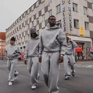 Puede incluir: Cuatro personas con ch&aacute;ndales grises a juego caminan por una calle de la ciudad. Cada persona lleva una sudadera con capucha y pantalones de ch&aacute;ndal holgados. El fondo incluye un edificio con las palabras "Hasbro House".