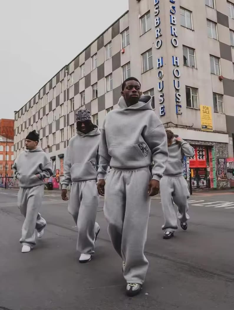 Puede incluir: Cuatro personas con ch&aacute;ndales grises a juego caminan por una calle de la ciudad. Cada persona lleva una sudadera con capucha y pantalones de ch&aacute;ndal holgados. El fondo incluye un edificio con las palabras "Hasbro House".
