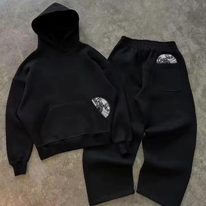 Puede incluir: Conjunto negro de sudadera con capucha y pantal&oacute;n deportivo a juego. La sudadera tiene un bolsillo delantero y una capucha. Ambas piezas presentan un gr&aacute;fico circular. El conjunto est&aacute; hecho de un material suave, ideal para uso casual.