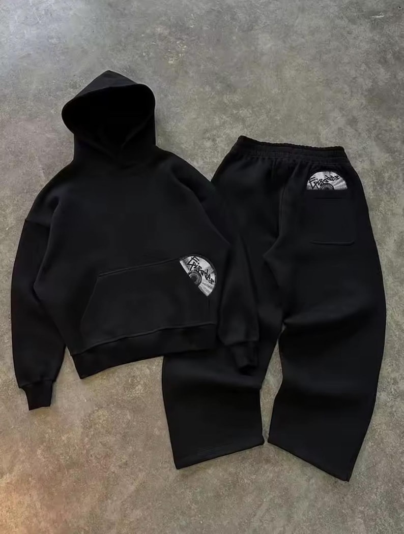 Puede incluir: Conjunto negro de sudadera con capucha y pantal&oacute;n deportivo a juego. La sudadera tiene un bolsillo delantero y una capucha. Ambas piezas presentan un gr&aacute;fico circular. El conjunto est&aacute; hecho de un material suave, ideal para uso casual.