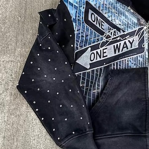 May include: Black zip-up hoodie with a cityscape graphic and "ONE WAY" text. The design features a blue and white cityscape with arrow signs. The left sleeve is embellished with small, round, silver-colored studs.