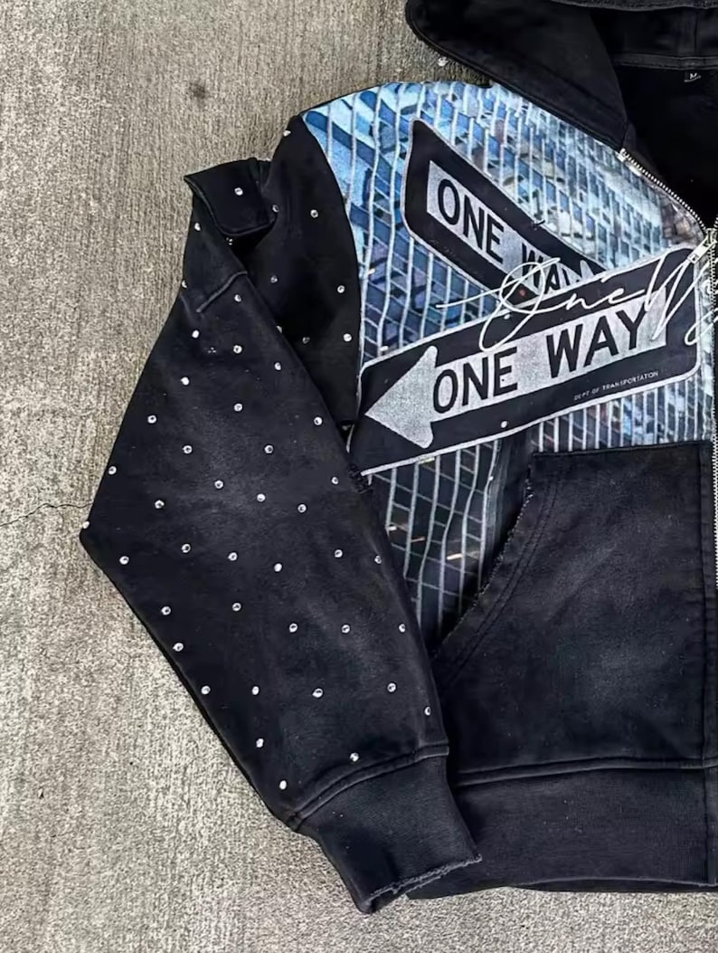 May include: Black zip-up hoodie with a cityscape graphic and "ONE WAY" text. The design features a blue and white cityscape with arrow signs. The left sleeve is embellished with small, round, silver-colored studs.