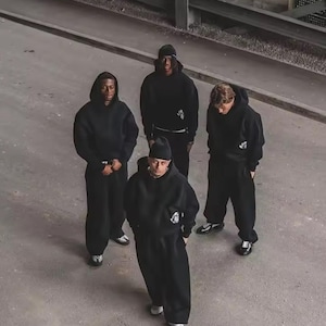 Puede incluir: Cuatro personas con sudaderas negras con capucha y pantalones de ch&aacute;ndal a juego de pie sobre una superficie de hormig&oacute;n gris. Cada persona lleva un gorro o capucha negra. El grupo lleva zapatillas blancas o negras. La imagen est&aacute; tomada desde un &aacute;ngulo alto.