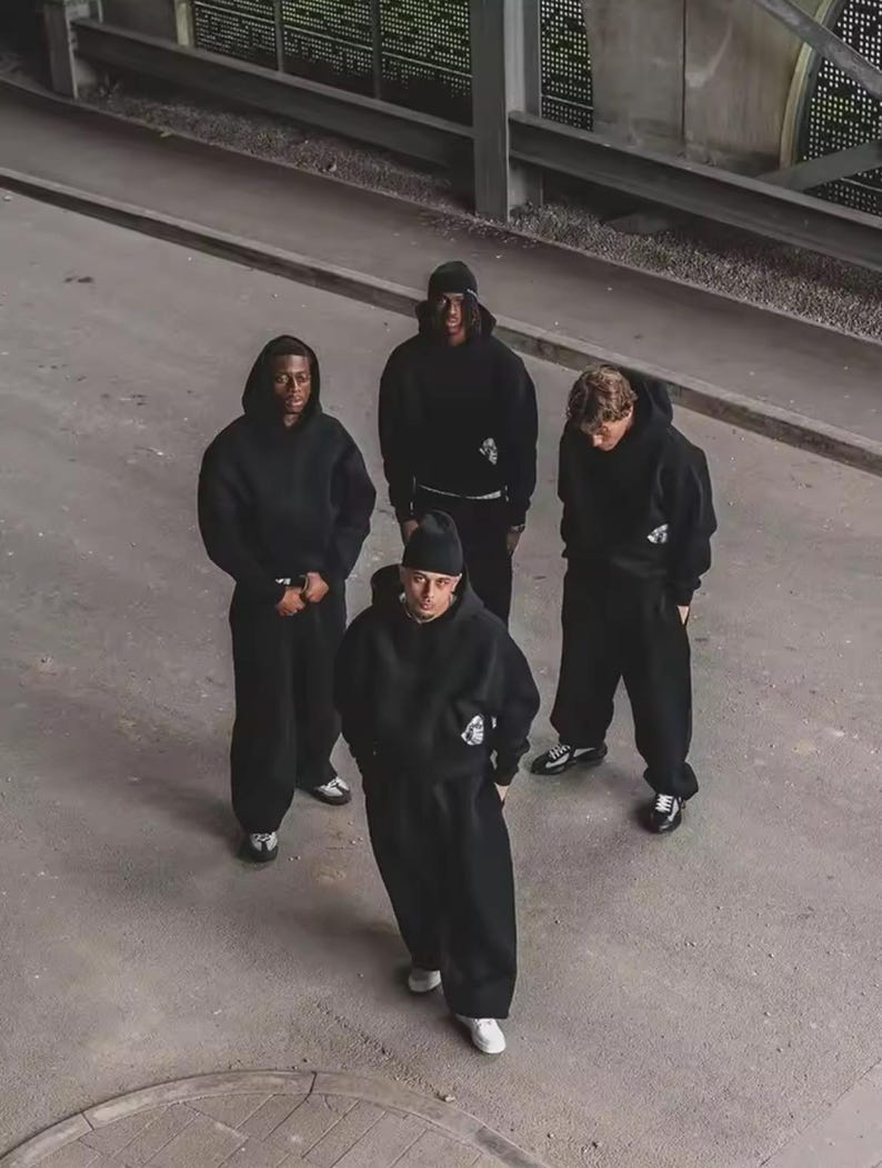 Puede incluir: Cuatro personas con sudaderas negras con capucha y pantalones de ch&aacute;ndal a juego de pie sobre una superficie de hormig&oacute;n gris. Cada persona lleva un gorro o capucha negra. El grupo lleva zapatillas blancas o negras. La imagen est&aacute; tomada desde un &aacute;ngulo alto.