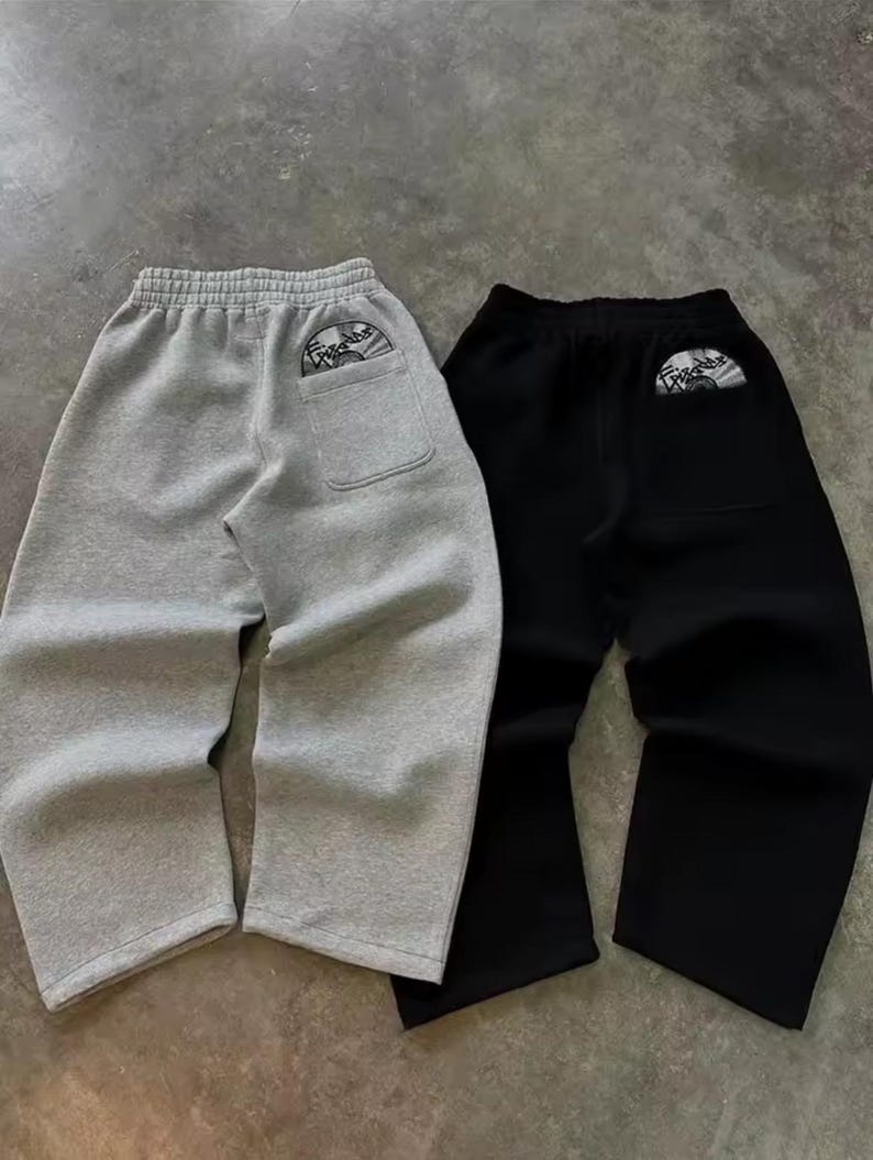 Puede incluir: Se muestran dos pares de pantalones de ch&aacute;ndal, uno gris y otro negro. Cada par tiene un bolsillo trasero con un peque&ntilde;o gr&aacute;fico circular. Los pantalones de ch&aacute;ndal tienen un corte holgado y una cintura con cord&oacute;n.