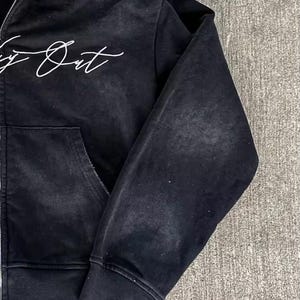 May include: Black zip-up hoodie with a distressed, vintage look. Features a white script design on the chest and a front pocket. The hoodie has a ribbed cuff and hem. The fabric appears to be a soft, worn material.