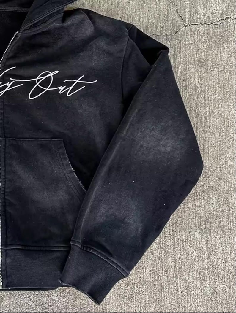 May include: Black zip-up hoodie with a distressed, vintage look. Features a white script design on the chest and a front pocket. The hoodie has a ribbed cuff and hem. The fabric appears to be a soft, worn material.