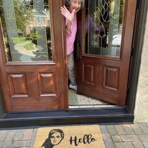 May include: A rectangular doormat made of natural fibers with a black illustration of a woman's face and the word "Hello" in black script. The mat is placed in front of a brown wooden door. An elderly woman waves from the doorway.