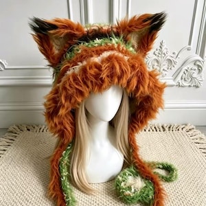 May include: A hooded hat featuring long, plush, rust-orange faux fur with prominent pointed ears. The interior and trim are crafted from a textured, variegated green and white yarn. Two long, furry earflaps extend downward from the main hood.