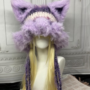 May include: A fuzzy, lavender-colored crochet hat featuring prominent, pointed cat-like ears. The hat is constructed with a thick, textured yarn and includes long, dangling braided ties in a deep purple shade, hanging down from the sides.