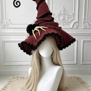 May include: A crocheted witch hat in deep burgundy with a black spiraled tip and black trim along the wide, wavy brim. A black fuzzy pom-pom is attached to the side, accented with pale yellow, multi-legged spider-like appendages.