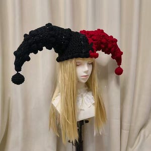 May include: A two-toned jester hat crafted from black and red yarn, featuring a textured bobble stitch pattern throughout. The hat has two long, curved points that extend outwards, each finished with a round pom-pom at the tip and a black lace-up detail.
