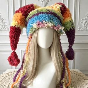 Op de afbeelding: Een veelkleurige narrenmuts gemaakt van getextureerd, ruig garen in de kleuren rood, oranje, geel, groen, blauw en paars. De muts heeft twee lange, puntige zijflappen die eindigen in pompons en lange, bungelende franjes aan de zijkanten.