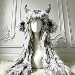 May include: A grey crochet hood featuring long faux fur trim, two pointed horns on top, and two circular ear-like accents on the sides. The hood is displayed on a white mannequin head, with long grey faux fur strands draping down the front.