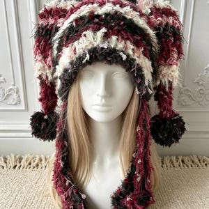 Könnte beinhalten: Eine mehrfarbige Narrenkappe mit einem strukturierten, flauschigen Strickmuster in den Farben Dunkelrot, Schwarz und Creme. Die Mütze verfügt über zwei lange, herabhängende Seitenteile, die mit dunklen Bommeln versehen sind.