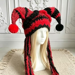 Puede incluir: Un gorro de estilo bufón con textura difusa y un patrón de rayas rojas y negras vibrantes. El gorro tiene dos extensiones laterales largas y puntiagudas rematadas con pompones y dos borlas trenzadas largas que cuelgan a los lados.