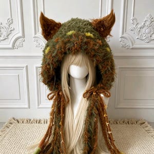 May include: A fuzzy, hooded headpiece featuring a textured blend of olive green and brown faux fur. The hood includes two prominent, pointed ears at the top and long, dangling braided ties on each side, accented with small yellow floral details.