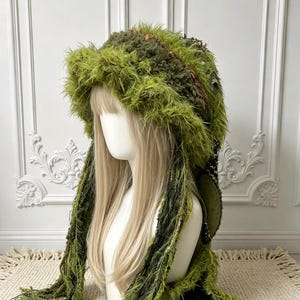 May include: A textured, moss-green crochet hood featuring long, shaggy fringe detailing. The headpiece is crafted from varied green yarn weights, creating a dense, layered appearance with extended side panels that drape down to the shoulders.