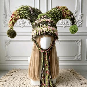 Pode incluir: Um gorro estilo bobo da corte feito de fio texturizado e felpudo em tons de verde-oliva, rosa claro e creme. O gorro apresenta dois chifres longos e curvos que se estendem pelas laterais, cada um com um pompom verde-oliva e laços trançados.