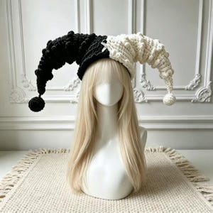 May include: A two-toned jester hat crafted from chunky yarn, featuring a black left side and a white right side. The hat has a textured, bobbled surface with long, drooping points on each side, each finished with a round pom-pom at the end.