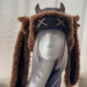 May include: A dark gray knit hat featuring two small brown horns on top and long, plush brown faux fur ear flaps. The front of the hat displays two embroidered gold-colored X symbols. The hat is displayed on a mannequin head with light hair.