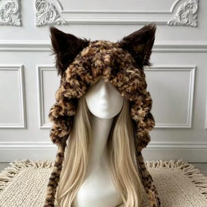 Può includere: Un cappuccio in pelliccia sintetica morbida con motivo leopardato marrone e nero. Il cappuccio presenta due orecchie da animale appuntite e prominenti di colore marrone scuro sulla parte superiore e due lunghi lacci abbinati ai lati.